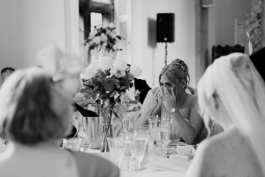 A close-up of a woman with blonde hair, visibly emotional, wiping her tears at a wedding reception, surrounded by guests and floral decorations.
