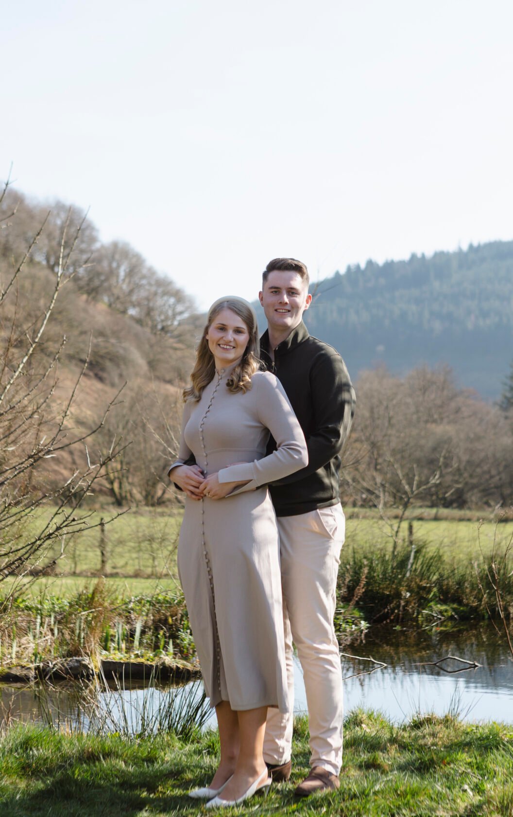 A couple stands together by a serene pond, with the woman in a light grey dress and the man in a black sweater. They are surrounded by nature and gently rolling hills in the background.