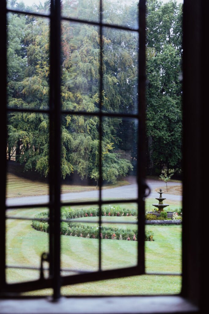 Looking through a window with a grid pattern, revealing a lush garden with a circular flower bed and a fountain surrounded by trees.