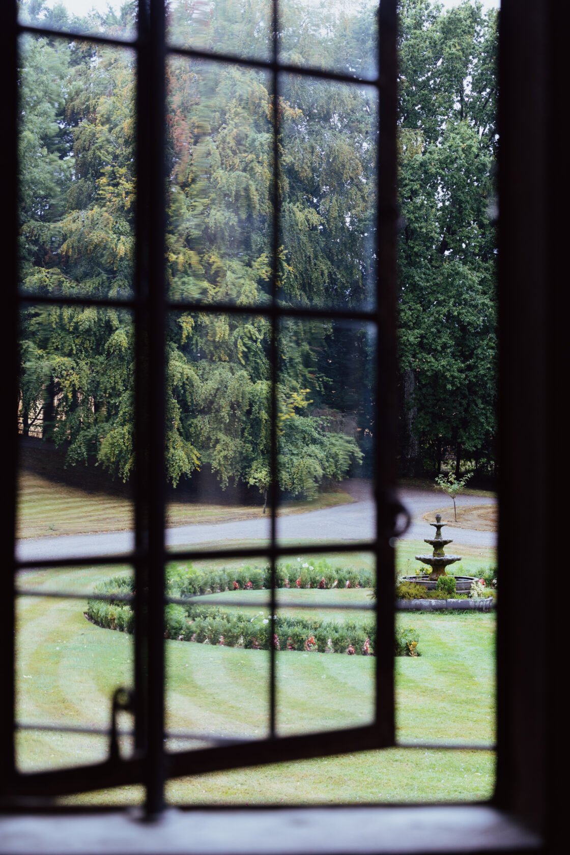 Looking through a window with a grid pattern, revealing a lush garden with a circular flower bed and a fountain surrounded by trees.