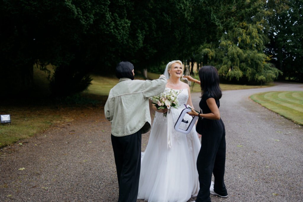 A smiling bride in a beautiful wedding dress holds a bouquet, surrounded by two people adjusting her veil and makeup in an outdoor setting.