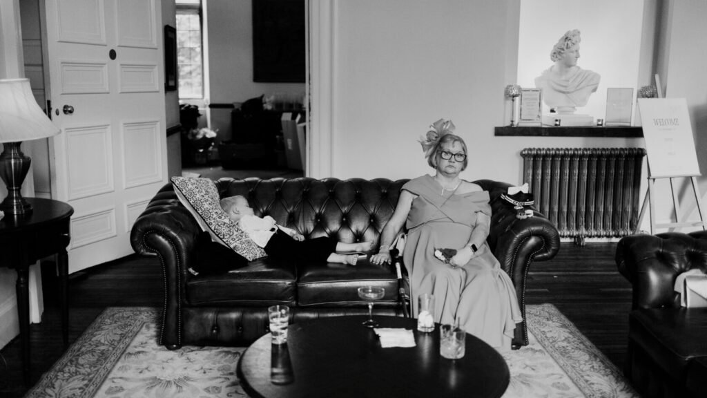 An elderly woman in a formal dress seated on a leather sofa, with a child asleep beside her, surrounded by a tastefully decorated room.