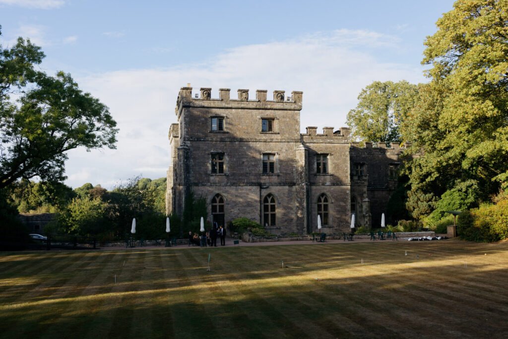 A historic stone castle with battlements, large windows, and a well-maintained lawn, surrounded by trees and outdoor seating areas.
