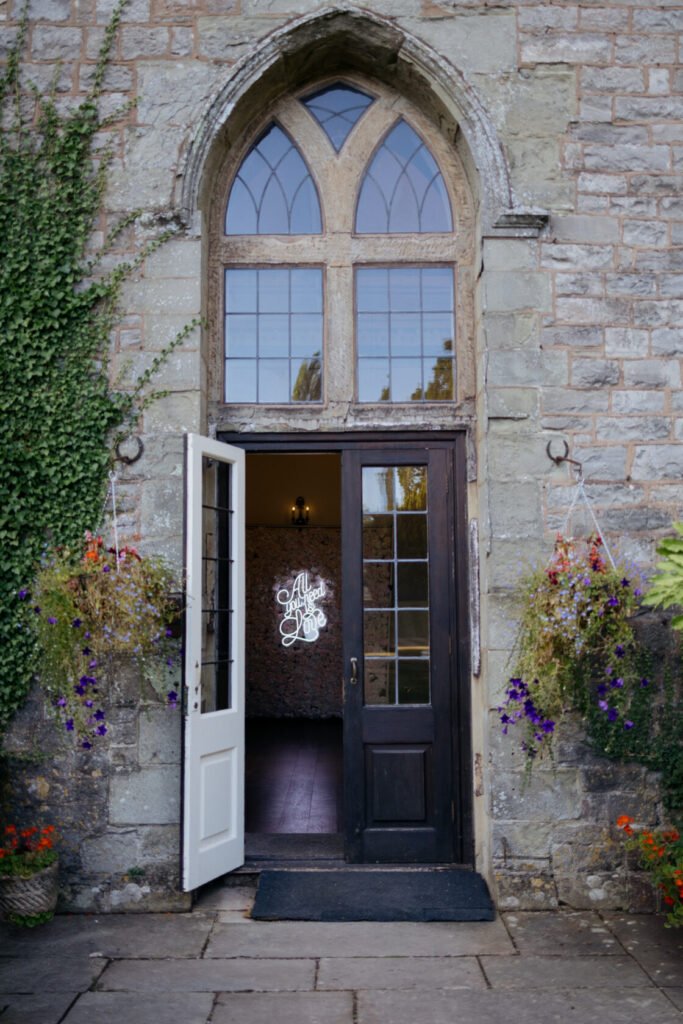 Elegant entrance of a stone building with an arched window and wooden doors, featuring hanging flower baskets and a neon sign saying 'All You Need is Love'.