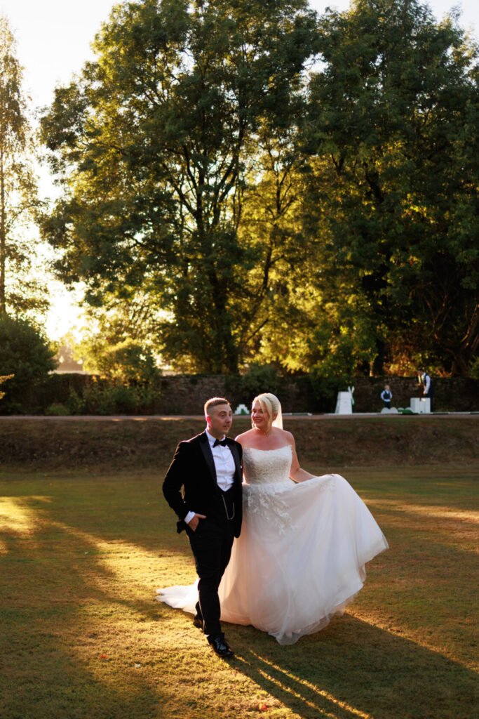 A bride in a white wedding gown and a groom in a black tuxedo stroll together in a sunlit outdoor setting, surrounded by trees.