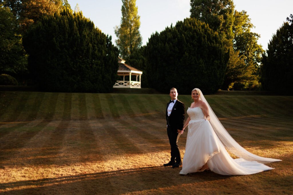 Bride and groom holding hands, walking on a grassy lawn with trees and a gazebo in the background during golden hour.
