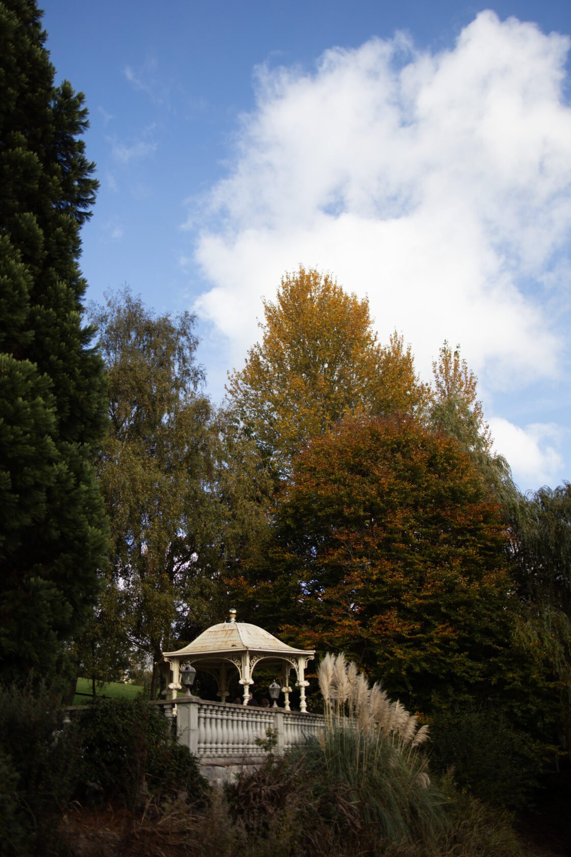 Scenic Autumn Gazebo Amidst Colourful Trees A decorative gazebo surrounded by vibrant autumn trees under a partly cloudy sky.